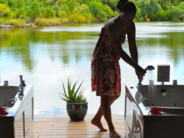 A person in a patterned dress adjusts a faucet near two tubs by a serene lake surrounded by lush greenery.