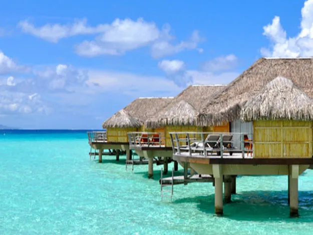 Overwater bungalows with thatched roofs sit above clear turquoise ocean water under a bright blue sky with fluffy clouds.