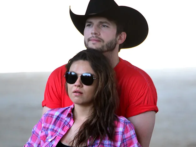 A couple outdoors, man in a cowboy hat and red shirt, woman in sunglasses and plaid shirt, standing closely.