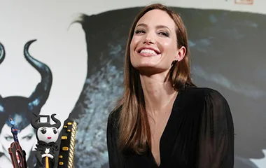 Angelina Jolie smiling, celebrating her 39th birthday in China with "Maleficent" backdrop and themed cake.