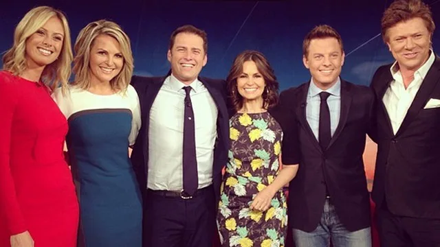 Group photo of six smiling TV hosts in formal attire on the set of a morning show.