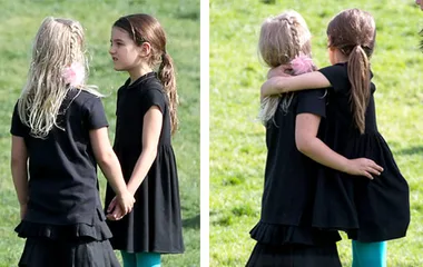 Two young girls in black dresses holding hands and hugging outdoors, showing friendship and affection.