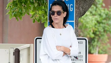 Sandra Bullock outside in casual attire, wearing sunglasses, standing near a tree with a handicapped parking sign behind her.