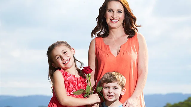 Woman in an orange dress smiling with two children, one holding a red rose, outside on a clear day.