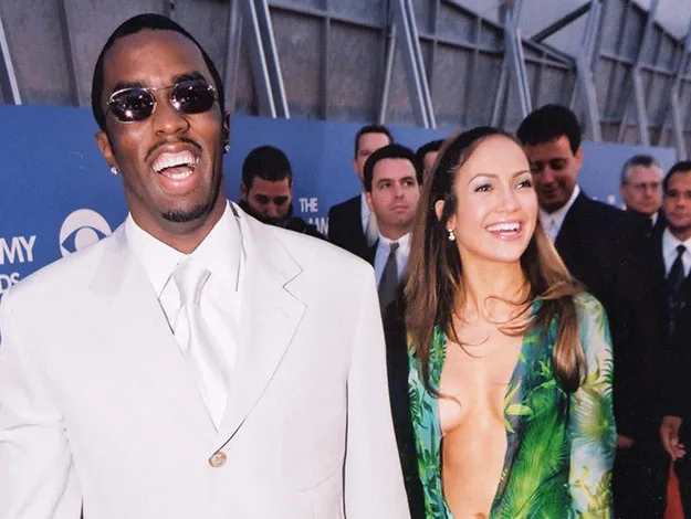 Two people smiling on a red carpet, wearing formal attire; a man in a suit and woman in a green patterned dress.