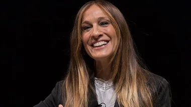 Sarah Jessica Parker smiles on stage, wearing a dark blazer and white lace top, hinting at a "Sex and the City" return.