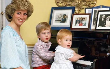 Princess Diana smiling with two young children playing at a piano, framed family photos in the background.