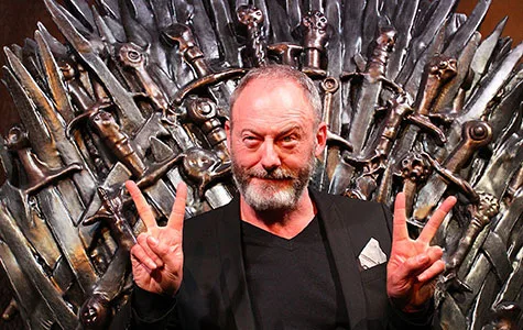 Man with gray beard posing with peace signs in front of the Iron Throne from Game of Thrones.