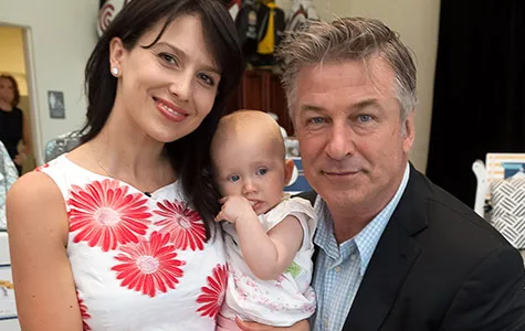 A couple with a baby, smiling together indoors. The woman is wearing a white dress with red flowers.
