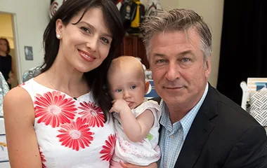 A couple with a baby, smiling together indoors. The woman is wearing a white dress with red flowers.