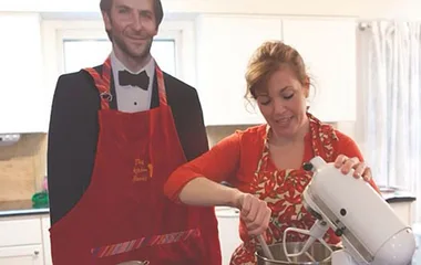 Cardboard cutout in tuxedo with apron beside woman using mixer in kitchen.