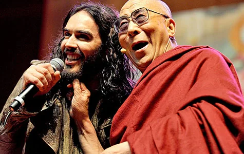 Two individuals smiling on stage, one holding a microphone, the other in red robes.