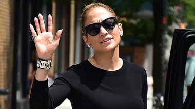 A person in sunglasses and a black top waves while stepping out of a vehicle, with buildings in the background.