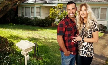 A couple smiling in front of a house, man in plaid shirt, woman in leopard print, lush garden in background.