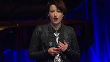 Singer Megan Washington speaks on stage, holding a phone, wearing a leather jacket against a dark, blurred background.