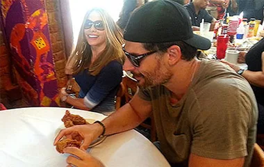 Celebrity couple having lunch at a restaurant, both wearing sunglasses and smiling, with other diners in the background.