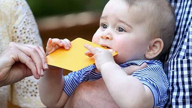 A baby in a striped shirt bites a yellow card while being held by an adult.