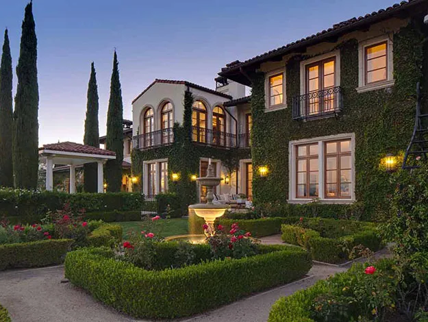 Mediterranean-style house with ivy-covered walls, large windows, and a fountain in a landscaped garden at dusk.