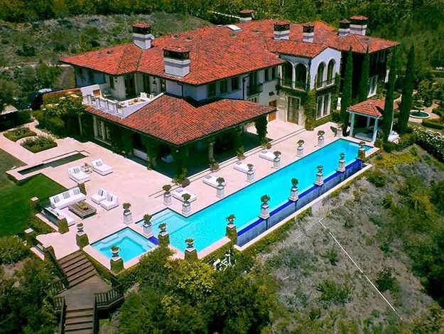 Luxury mansion with red-tiled roof, spacious patio, and large rectangular swimming pool surrounded by trees.