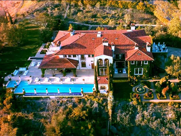Aerial view of a large, luxurious mansion with red roof tiles, pool, and surrounding gardens.