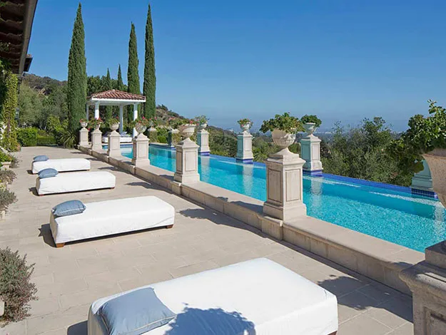 Luxurious outdoor pool with lined daybeds, surrounded by greenery and columns, under a clear blue sky.