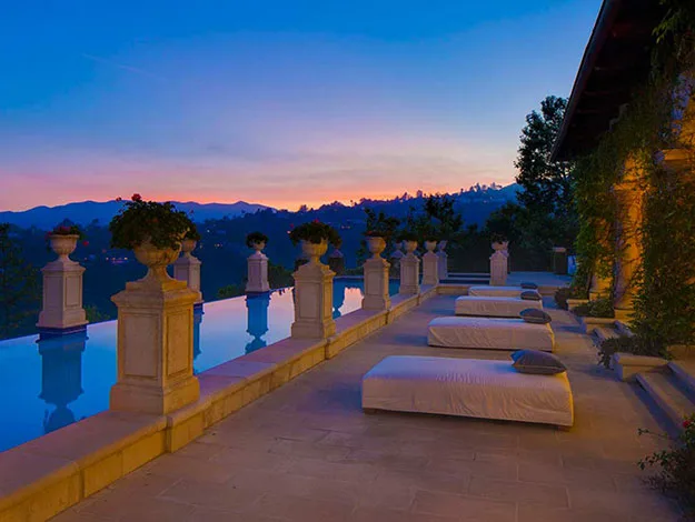 A serene infinity pool at sunset with lounge chairs and ornate columns, overlooking a distant mountain range.