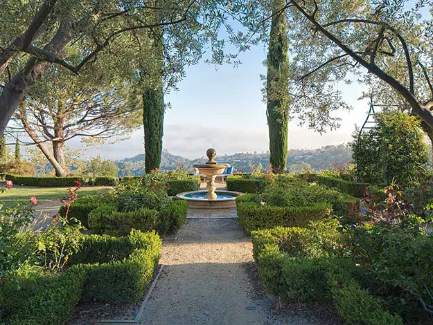 A picturesque garden with a central fountain, surrounded by trees and hedges, overlooking distant hills under a clear sky.