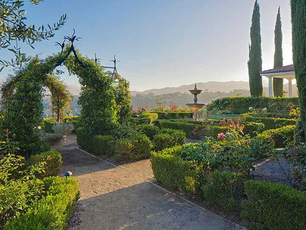 Beautiful garden with hedges, rose bushes, an archway covered in greenery, a central fountain, and tall cypress trees.