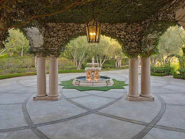 Outdoor pavilion with ivy-covered ceiling, columns, and a central decorative fountain surrounded by trees.