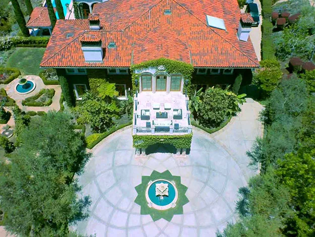 Aerial view of a large house with a red-tiled roof, circular driveway, and central fountain surrounded by greenery.