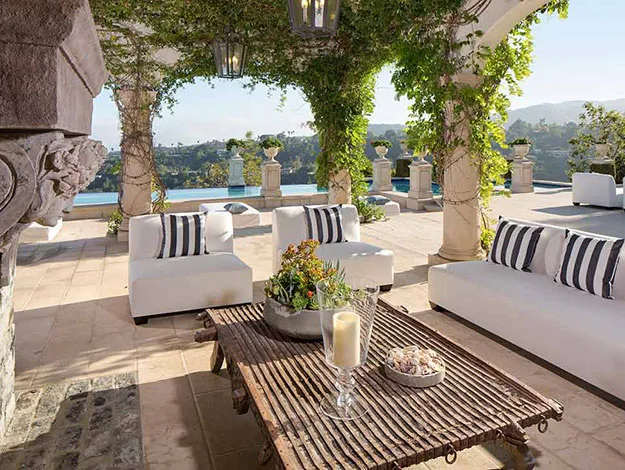 Outdoor patio with white sofas, striped cushions, vine-covered pergola, wooden table, candle, and distant hills view.