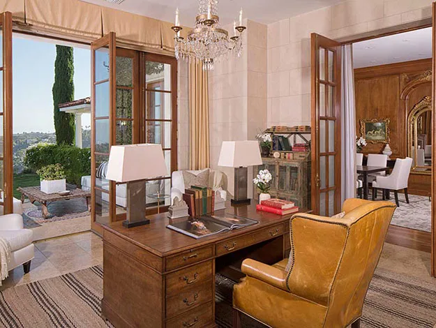 Office interior with wooden desk, leather chair, chandelier, open doors to a patio, and distant landscape view.