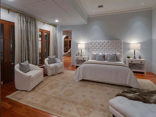 Luxurious bedroom with a tufted headboard, elegant chairs, soft lighting, and large windows with drapes.