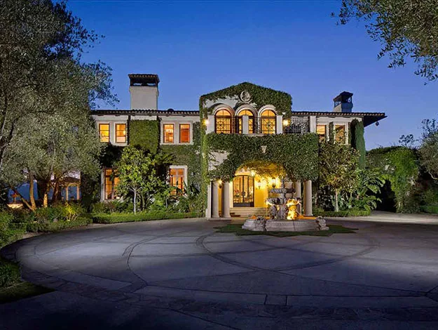 Luxurious estate with ivy-covered walls, illuminated windows, and a central fountain, under a deep blue evening sky.
