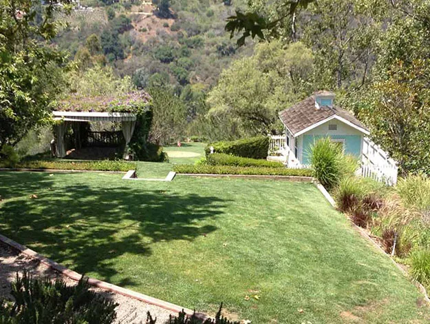 A lush garden with manicured lawn, small white cottage, and gazebo with mountains in the background.