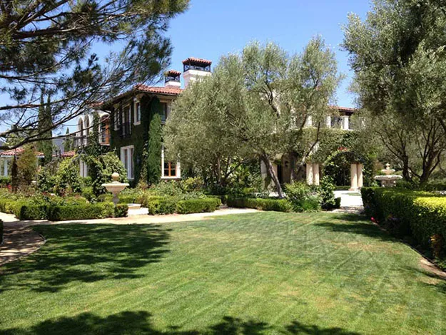 Elegant mansion with manicured lawn, tall trees, and lush garden under a clear blue sky.