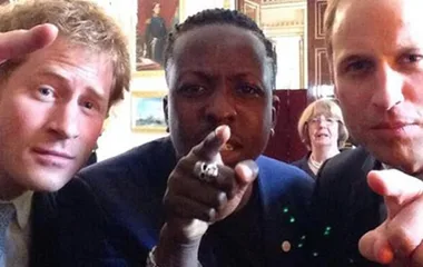 Three people taking a selfie, two pointing at the camera in a formal setting with paintings in the background.