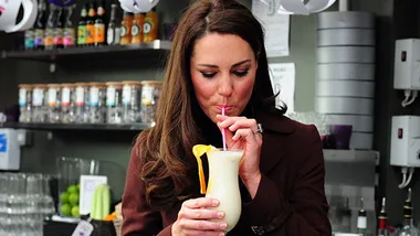 Woman sipping a creamy smoothie from a glass with a pink straw, surrounded by shelves of bottles and glasses.