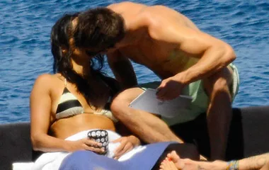 Couple kissing on a boat, surrounded by blue water, with the woman in a bikini and the shirtless man leaning over.