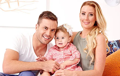 A smiling family with a man, woman, and a baby girl in a floral dress sitting together on a couch.