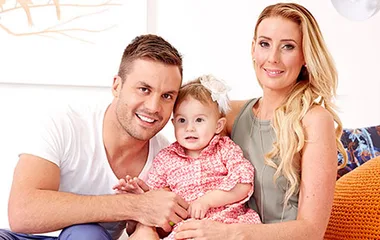 A smiling family with a man, woman, and a baby girl in a floral dress sitting together on a couch.