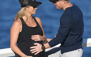 A couple outdoors, the woman wearing a cap and tank top shows her baby bump, while the man touches it gently.