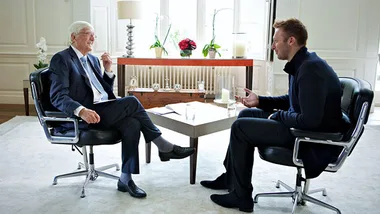 Two men engaged in a conversation, sitting in modern chairs facing each other in a well-lit, elegantly furnished room.