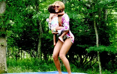 Woman in a floral swimsuit, holding a child, smiling in a lush green forest setting.