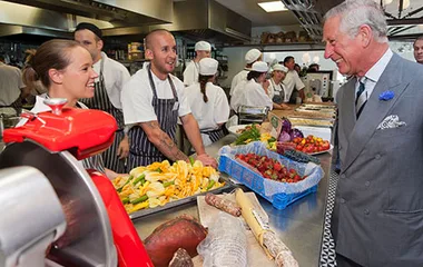 Prince Charles visits a restaurant kitchen, engaging with chefs and observing an array of fresh produce.