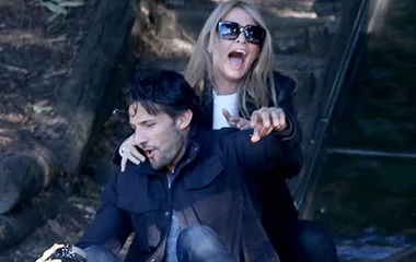 Couple laughing while sliding down together in an outdoor playground, both wearing sunglasses and casual clothing.