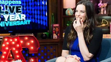 Liv Tyler laughs on a talk show set, seated in a blue dress, with colorful decor and a neon sign in the background.
