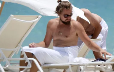 "Man with sunglasses and a bun sits on a beach chair, wrapped in a towel, near the ocean on a sunny day."