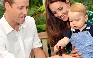 A family with a young child interacting with a butterfly on a bench in a garden setting.