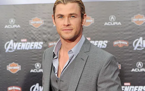 A man in a grey suit at a movie premiere event with "Avengers" logos in the background.